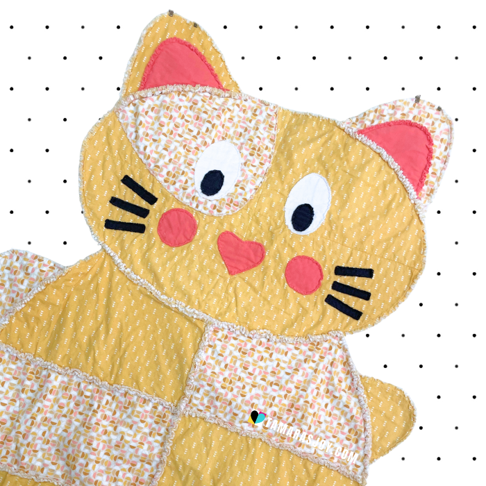 Yellow cat-shaped quilt with pink ears and cheeks on a white background
