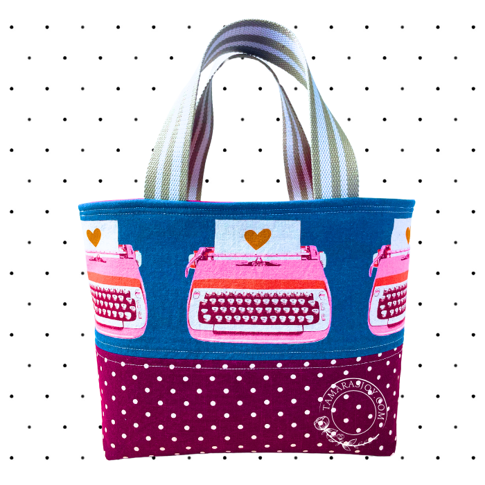 A multicolored small tote bag with a pattern of typewriters and hearts on a blue background, with a purple polka-dotted bottom panel and striped straps.