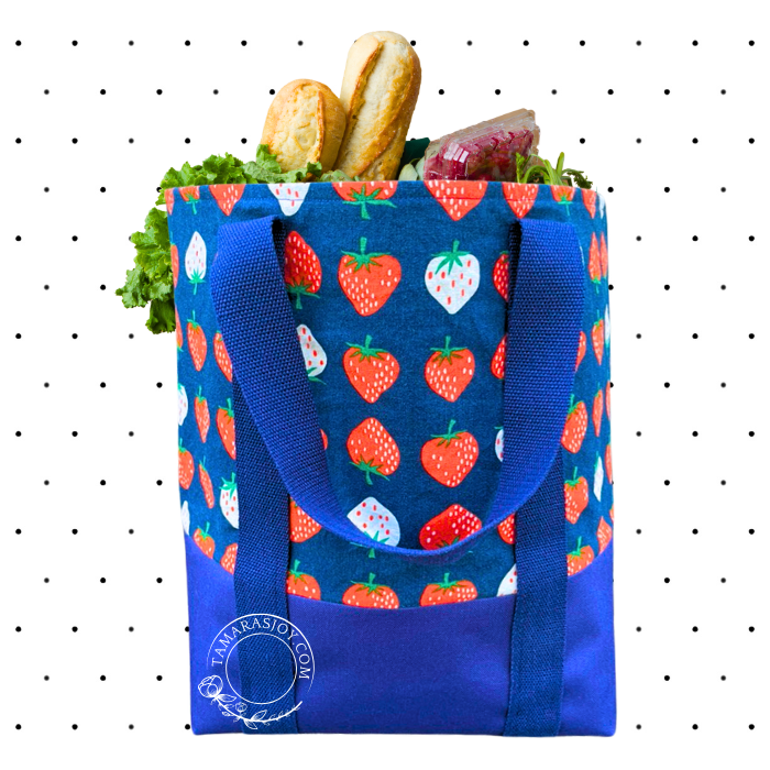A blue grocery tote bag with a pattern of strawberries, filled with groceries such as bread and vegetables.